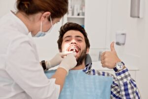 Young man giving thumbs-up while having mouth examined in dentist's chair. 